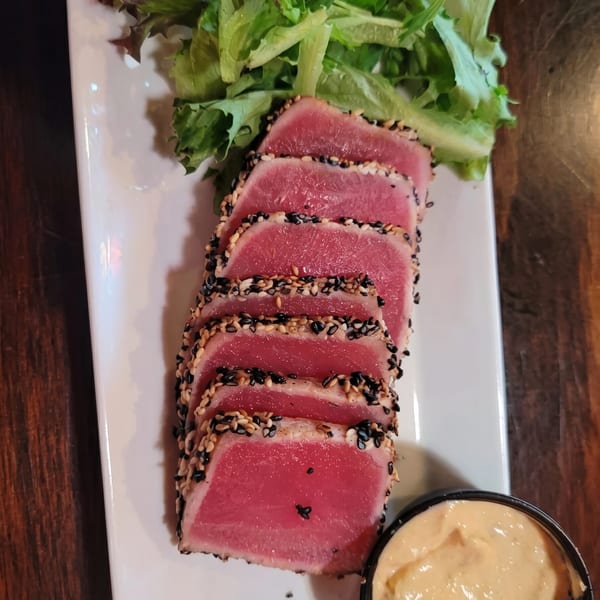 Seared tuna slices with sesame crust, fresh greens, and a side of creamy sauce on a white plate.
