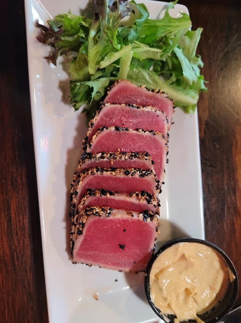 Seared tuna slices with sesame crust, fresh greens, and a side of creamy sauce on a white plate.
