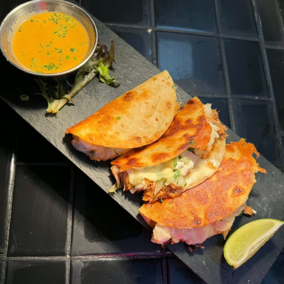 Three cheesy birria tacos with a side of consommé and a lime wedge on a dark slate.